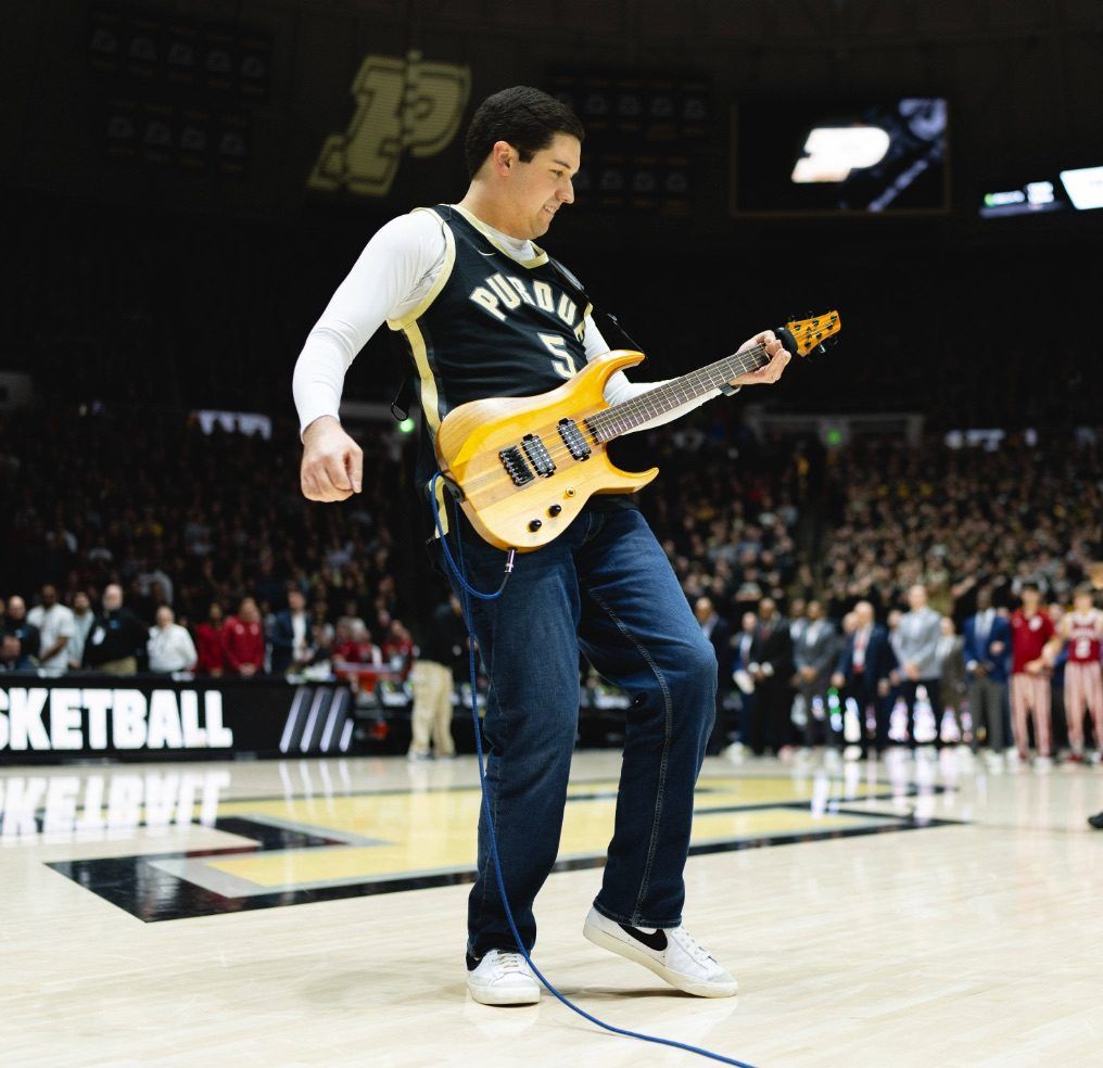 Noah Scott in a Purdue jersey playing the natural-wood baritone guitar on the Mackey Arena court
